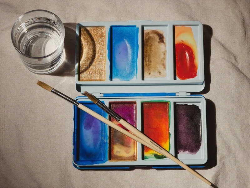 Offene Aquarellpalette mit leuchtenden Pigmenten, zwei Naturhaarpinseln und einem Glas klarem Wasser